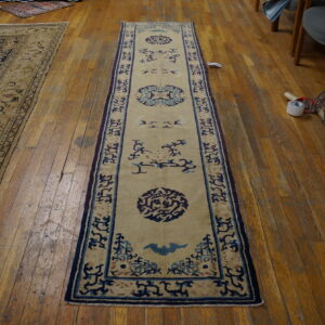 A low-pile beige runner rug features navy and light blue traditional motifs on wood plank flooring.