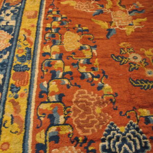 Close view of a high pile orange rug featuring navy and yellow floral designs and multi-banded striped borders.