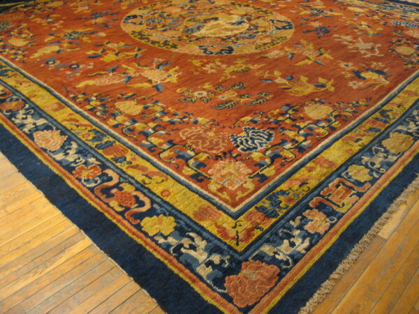 Rust-colored rug with complex blue, yellow, and salmon floral motifs and multi-banded borders lying on light wood flooring.