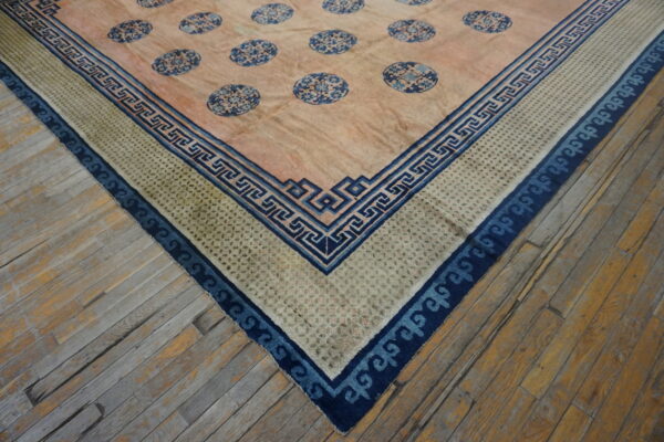 Corner view of a rug featuring a pink field, blue medallions, and intricate greek key borders on wood floor.