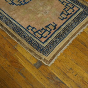 Low-pile pink and tan rug with a navy blue labyrinth border sits on diagonal distressed wood plank flooring.