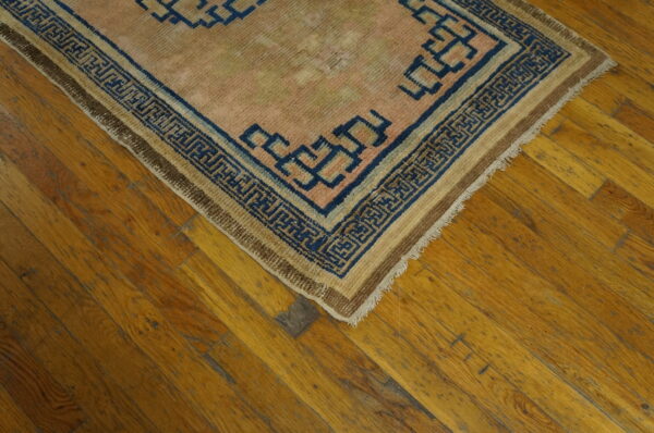 Low-pile pink and tan rug with a navy blue labyrinth border sits on diagonal distressed wood plank flooring.
