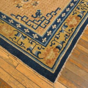 Corner of a low-pile tan rug with detailed blue and gold borders rests on light brown wood flooring.