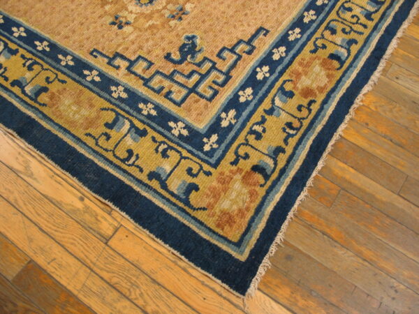 Corner of a low-pile tan rug with detailed blue and gold borders rests on light brown wood flooring.