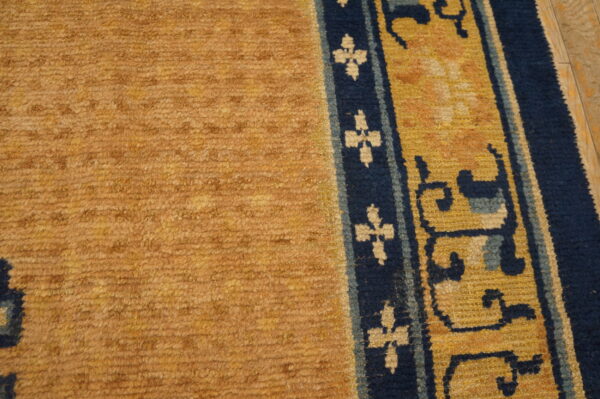 Textured gold rug pile with a dark blue and yellow ornamental border rests on light wooden flooring.