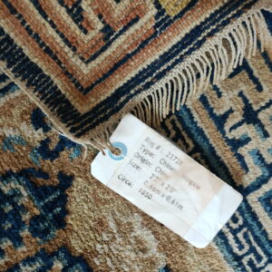 Low-pile rug fragment in navy, terracotta, and cream, showing fringes and an attached identification tag.