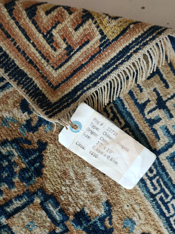 Low-pile rug fragment in navy, terracotta, and cream, showing fringes and an attached identification tag.