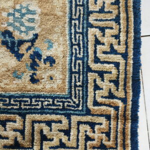 Textured tan and dark blue rug corner with floral and geometric patterns resting on white tiled flooring.