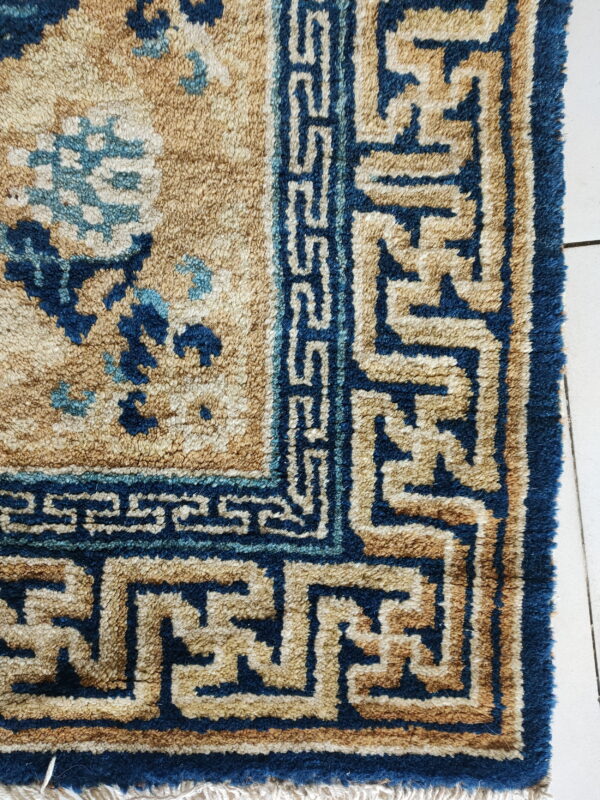 Textured tan and dark blue rug corner with floral and geometric patterns resting on white tiled flooring.