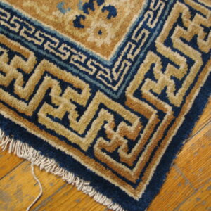 Deep blue and tan geometric border rug corner with fringe on worn wooden plank flooring.