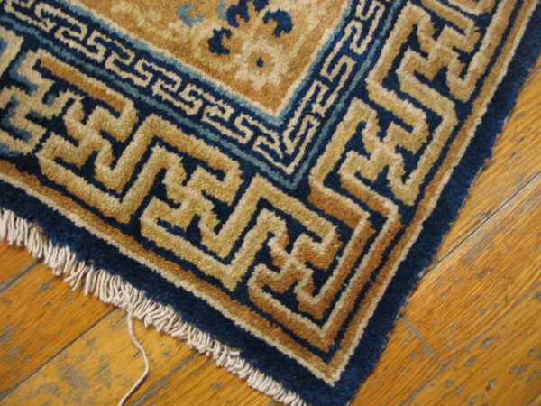 Deep blue and tan geometric border rug corner with fringe on worn wooden plank flooring.