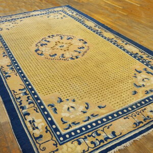 Tan rug with a dense dotted field and blue decorative borders lying on wide wooden planks.