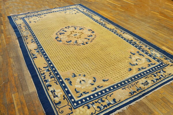 Tan rug with a dense dotted field and blue decorative borders lying on wide wooden planks.