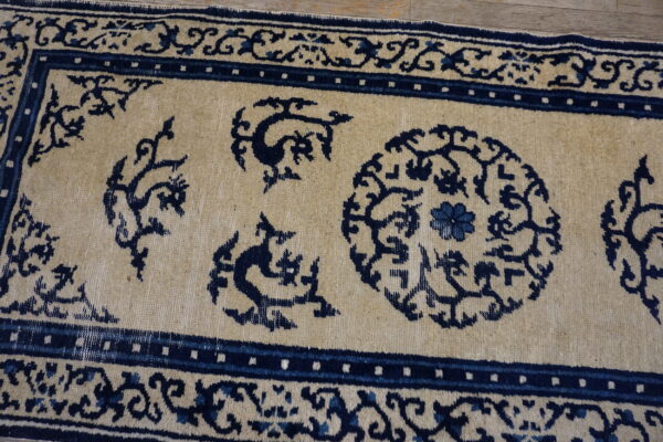 Close view of a tan and dark blue runner rug featuring dense patterns, lying on light wood flooring.