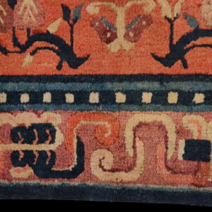A detailed view of a rug with a reddish field, dark blue vine motifs, and elaborate borders featuring cream dots and swirling blue bands.