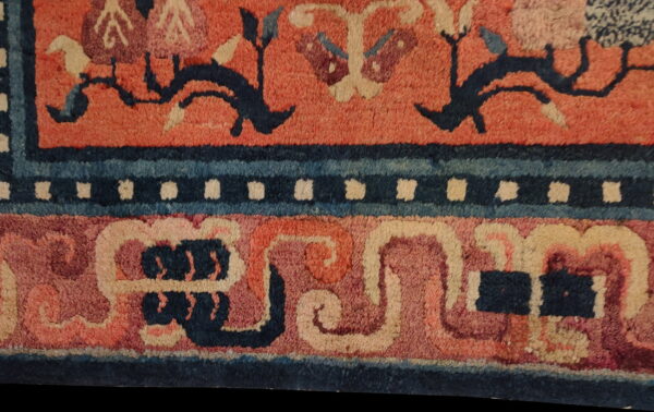 A detailed view of a rug with a reddish field, dark blue vine motifs, and elaborate borders featuring cream dots and swirling blue bands.