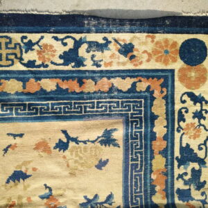 Cream rug with complex blue and rust borders, including floral scrollwork and a key pattern, resting on concrete.