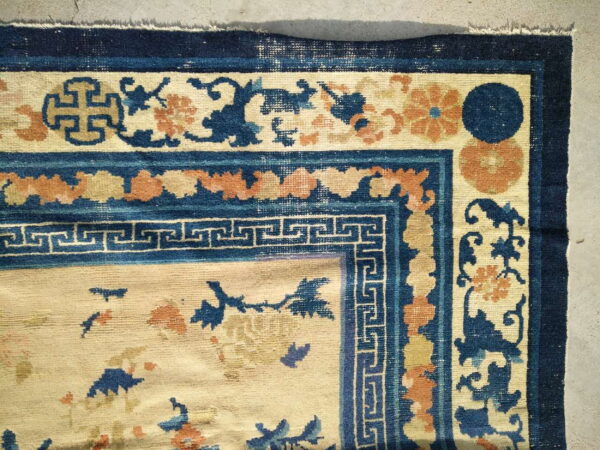 Cream rug with complex blue and rust borders, including floral scrollwork and a key pattern, resting on concrete.