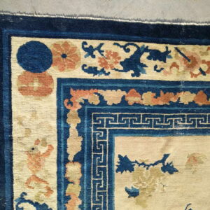 Beige field rug corner featuring navy blue borders, a geometric key pattern, and orange floral motifs.