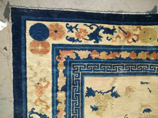 Beige field rug corner featuring navy blue borders, a geometric key pattern, and orange floral motifs.