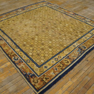 Square gold rug with geometric lattice field and blue, rust floral borders sits on a distressed wood floor.