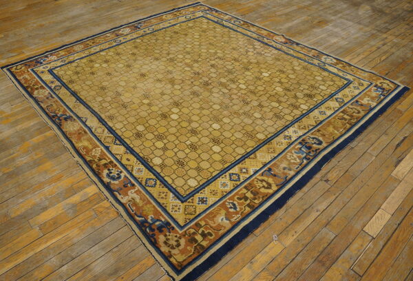 Square gold rug with geometric lattice field and blue, rust floral borders sits on a distressed wood floor.