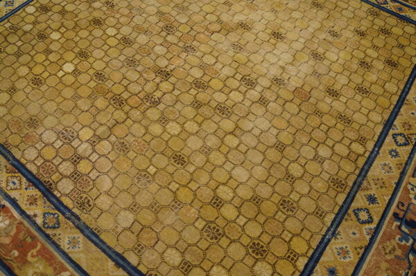 An aged yellow rug features a dense geometric lattice pattern outlined in brown, bordered by blue and terracotta stripes.