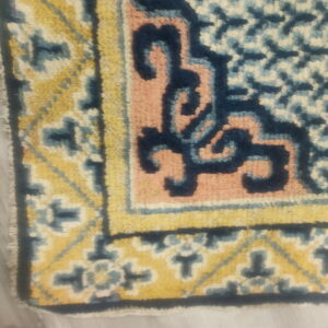 Ornate rug corner featuring geometric patterns in yellow, navy, light blue, and pink against light gray flooring.