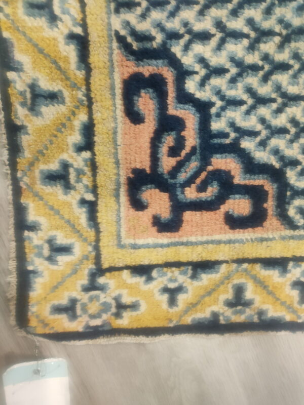 Ornate rug corner featuring geometric patterns in yellow, navy, light blue, and pink against light gray flooring.