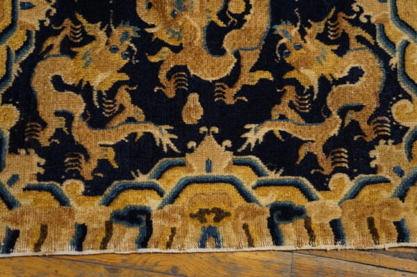 Intricate high-pile rug featuring gold and teal mythological creatures on a dark blue field over wood flooring.