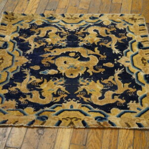 Square gold and navy rug with a stylized dragon motif resting on distressed wood planks.