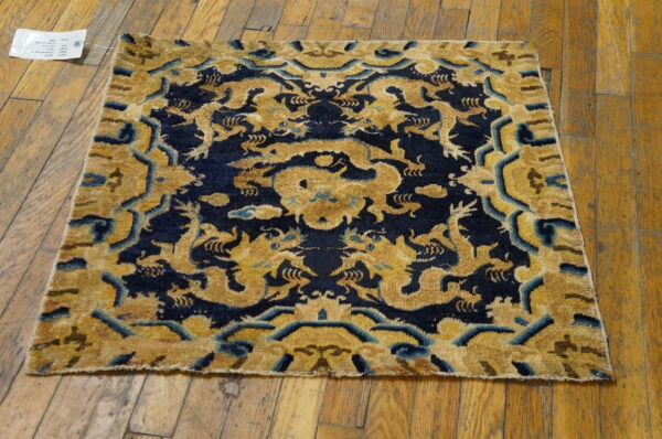 Square gold and navy rug with a stylized dragon motif resting on distressed wood planks.