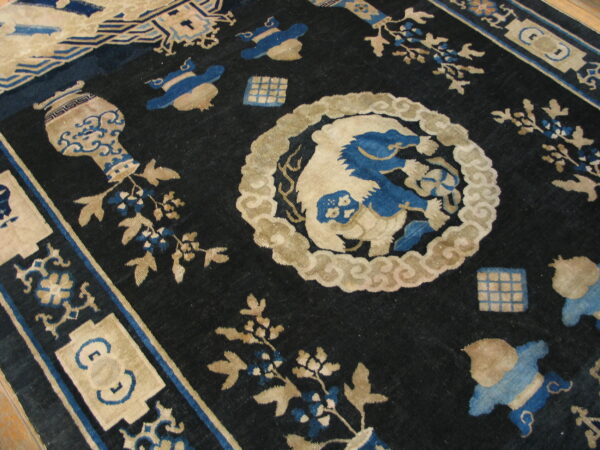 Ornate indigo rug featuring tan and blue mythological creatures, vases, and floral elements on a dark field.