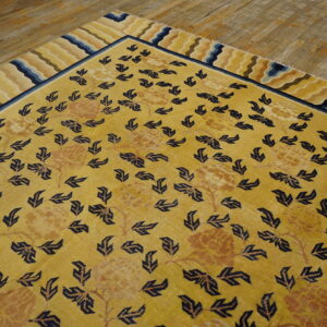 Gold rug featuring navy leaves, faded florals, and a wavy striped border rests on worn wood flooring.