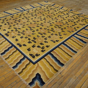 Large yellow rug with navy botanical patterns and wide, wavy striped borders on worn wood flooring.