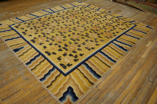 Golden yellow rug with blue foliage and a striped wavy border lays diagonally on light brown wood flooring.