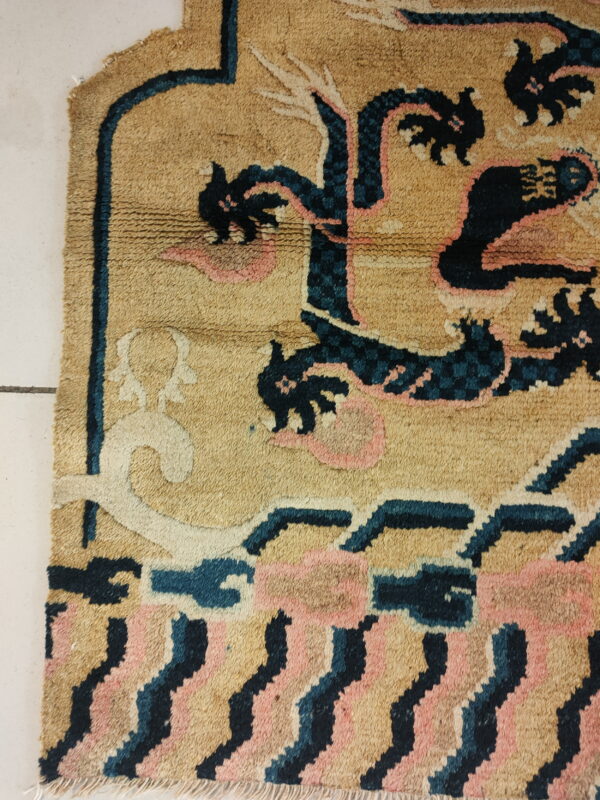 Detail of a tan rug with blue and pink stylized designs, showing fringe on a white tiled floor.