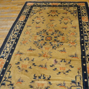 Muted gold rug with stylized blue and coral accents and a dark border lies on distressed wooden flooring.
