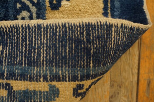 Close-up of a blue and beige patterned rug curled to show the warp and weft backing against wood planks.