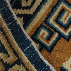 Close-up of a folded, textured rug displaying a dense geometric pattern in ochre, cream, and dark blue.
