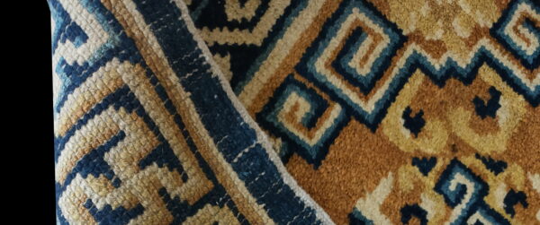Close-up of a folded, textured rug displaying a dense geometric pattern in ochre, cream, and dark blue.
