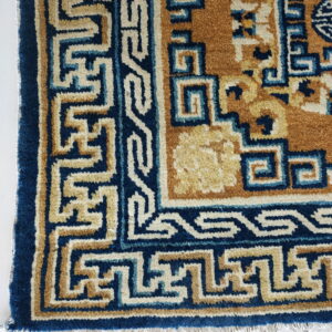 Blue, tan, and cream rug border featuring interlocking key patterns lying on a white tile floor with fringe.