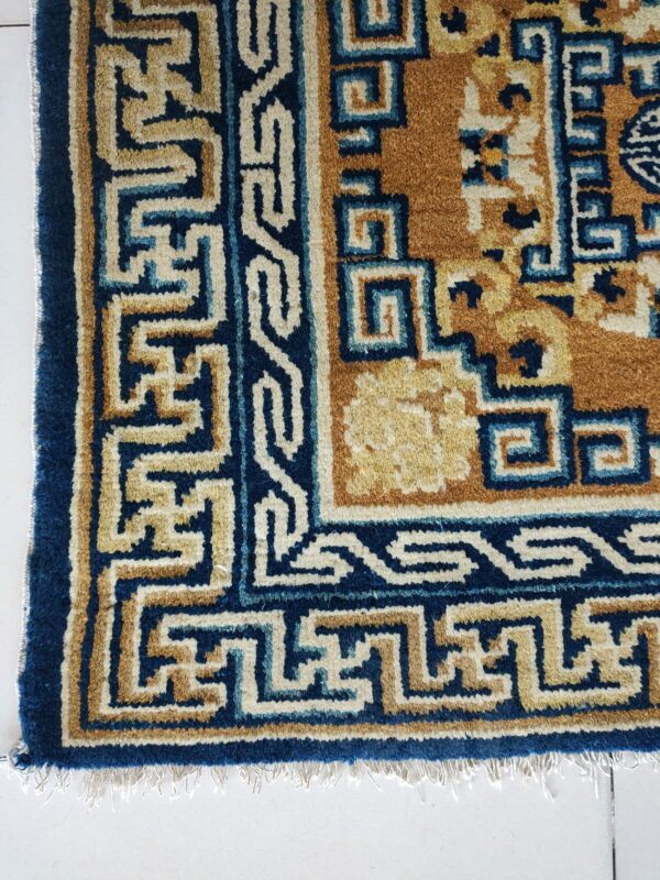 Blue, tan, and cream rug border featuring interlocking key patterns lying on a white tile floor with fringe.
