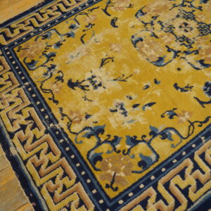 Mustard yellow rug with navy and tan floral and fret patterns on distressed wooden floor.