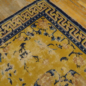 Distressed gold rug with navy blue floral patterns and geometric borders rests on light wood planks.