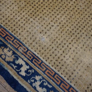 Worn low-pile rug with a spotted tan field and complex blue and peach border on wood.