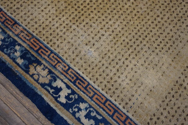 Worn low-pile rug with a spotted tan field and complex blue and peach border on wood.