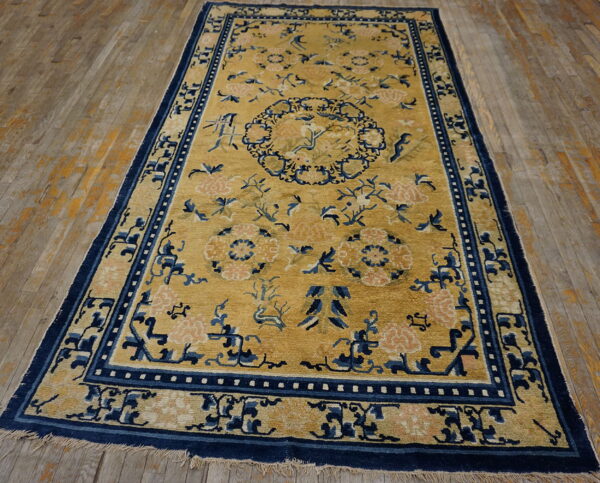 Gold rug with navy borders and pink floral patterns lies on light brown distressed wood planks.