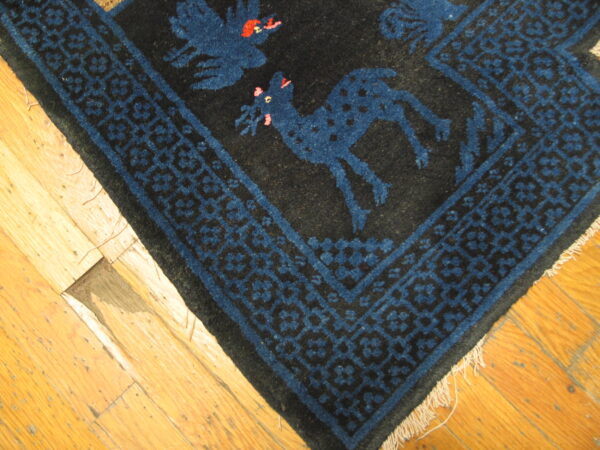 Close-up of a dark rug featuring blue animal and bird motifs, framed by a geometric border on hardwood flooring.