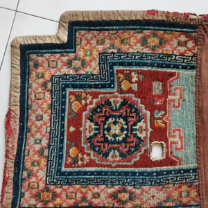 Worn red, blue, and teal patterned rug with a stepped corner and a hole, displayed on white tile.